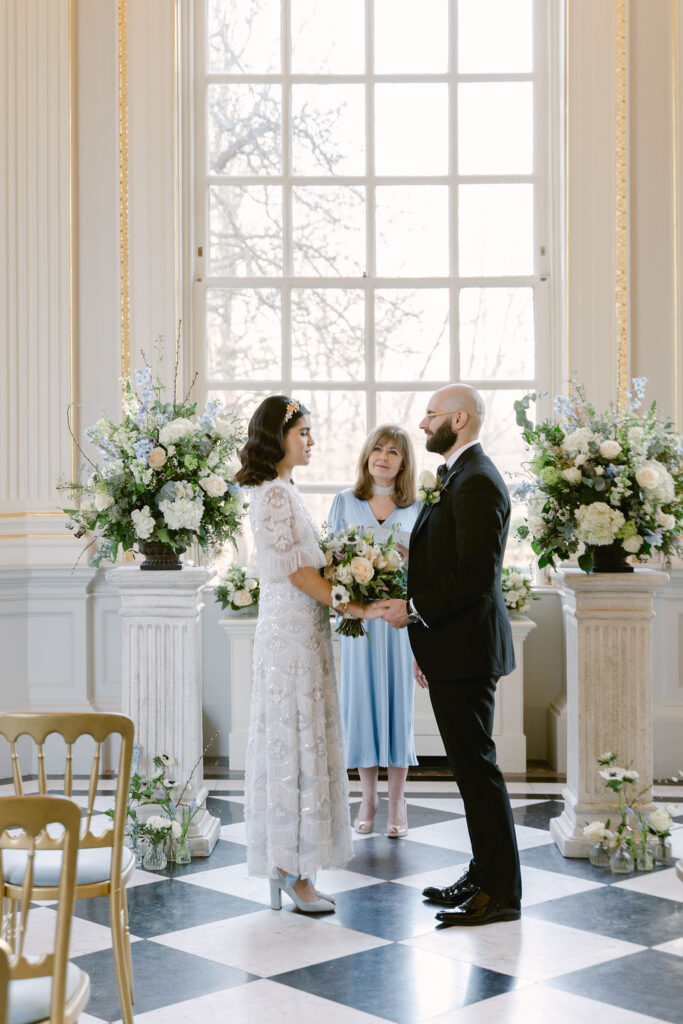 The bride and groom are partaking in a ceremony with the celebrant, who is dressed in a midi blue dress, with a large window in the background.