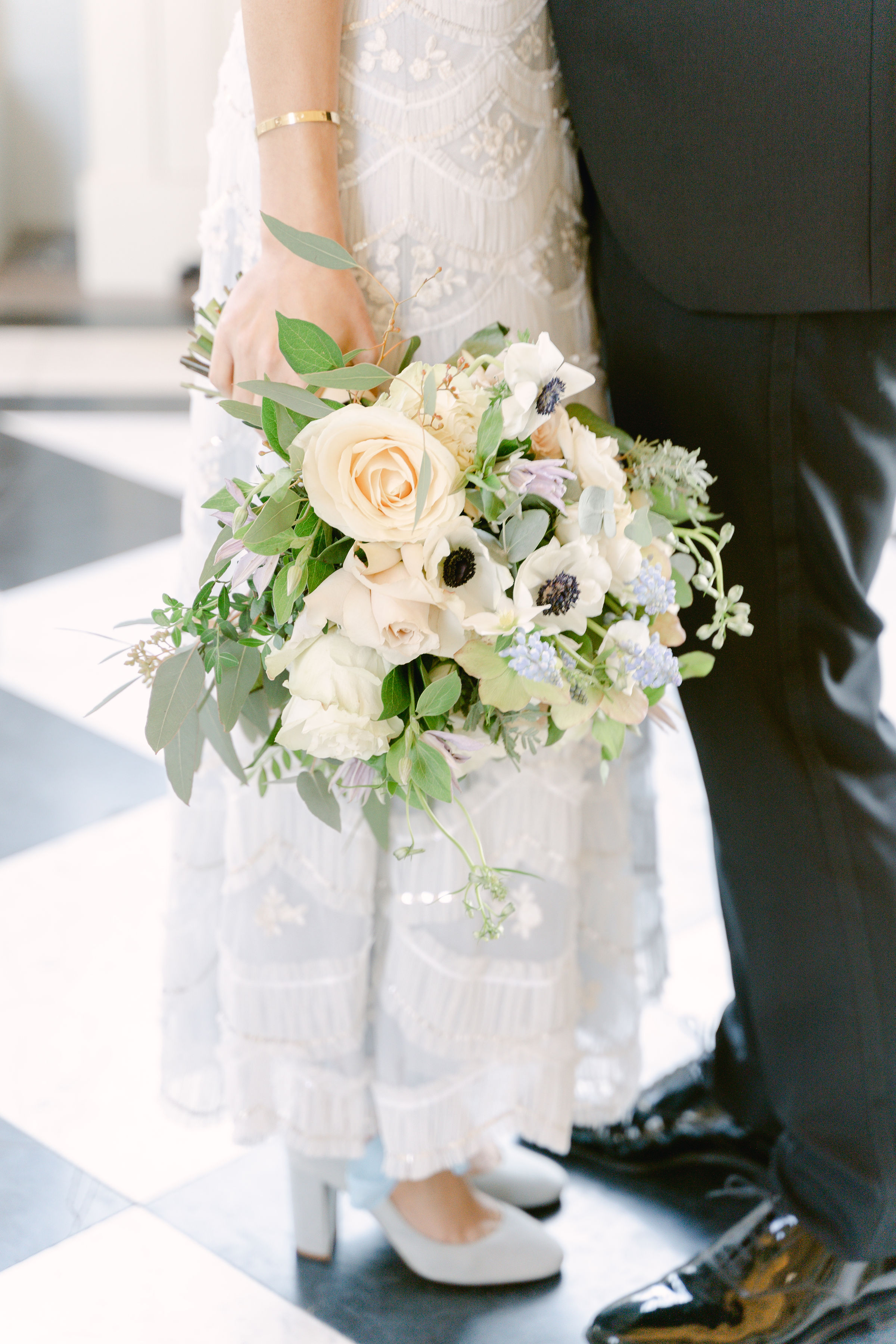 The bride holding her bouquet featuring anemones, roses, eucalyptus, hellebores and muscari. She is wearing a sparkly pale blue wedding gown and pale blue block heels.