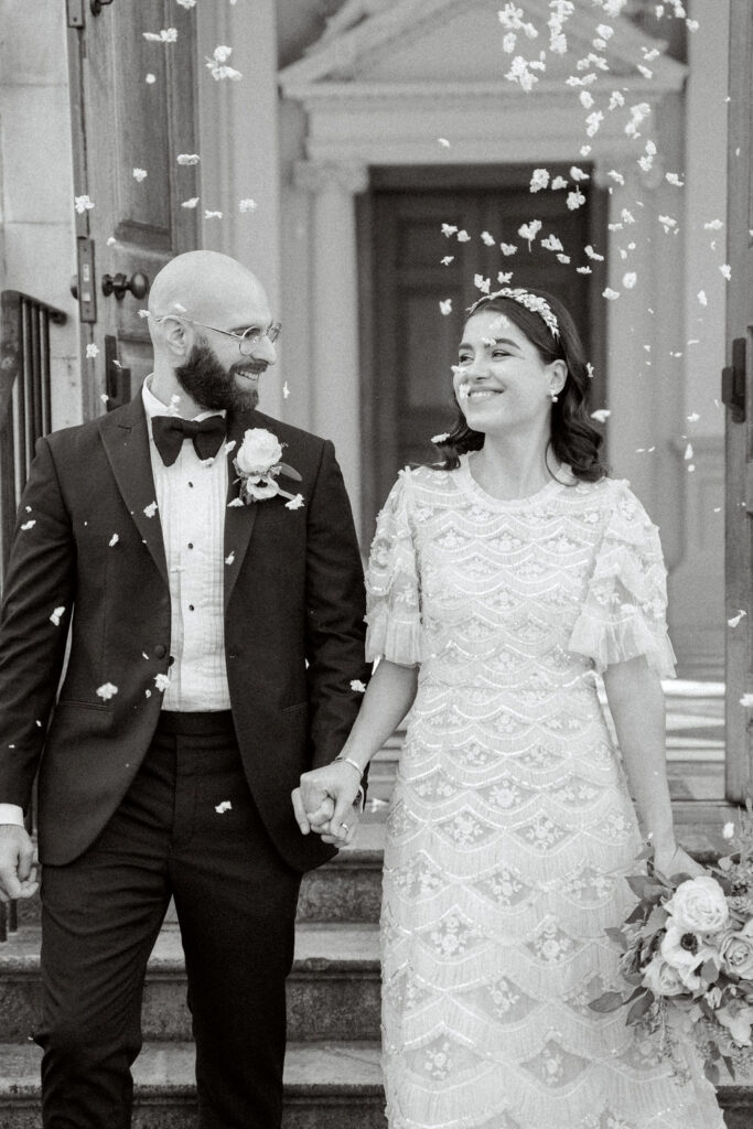 A black and white portrait of the bride and groom being showered in confetti following the fine art wedding ceremony.