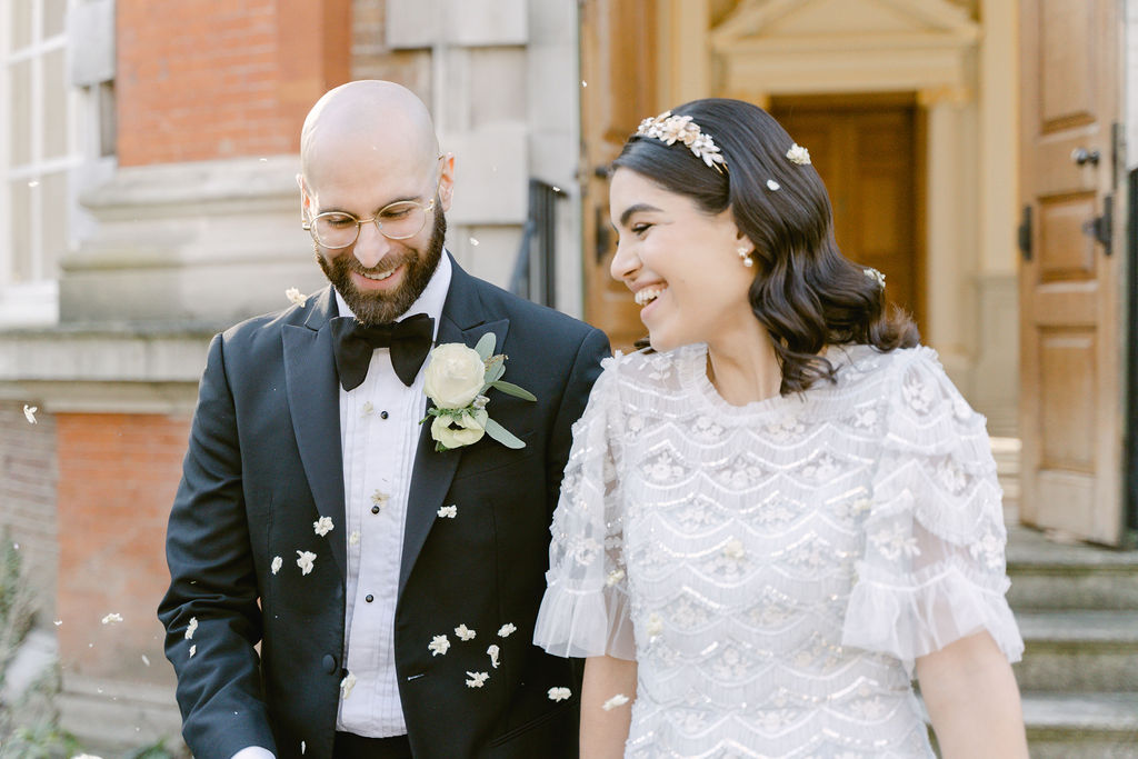 The bride and groom exiting the Octagon Room to the outside of the building, being showered in confetti. The groom is wearing a tuxedo with a buttonhole. The bride is smiling in a pale blue gown.