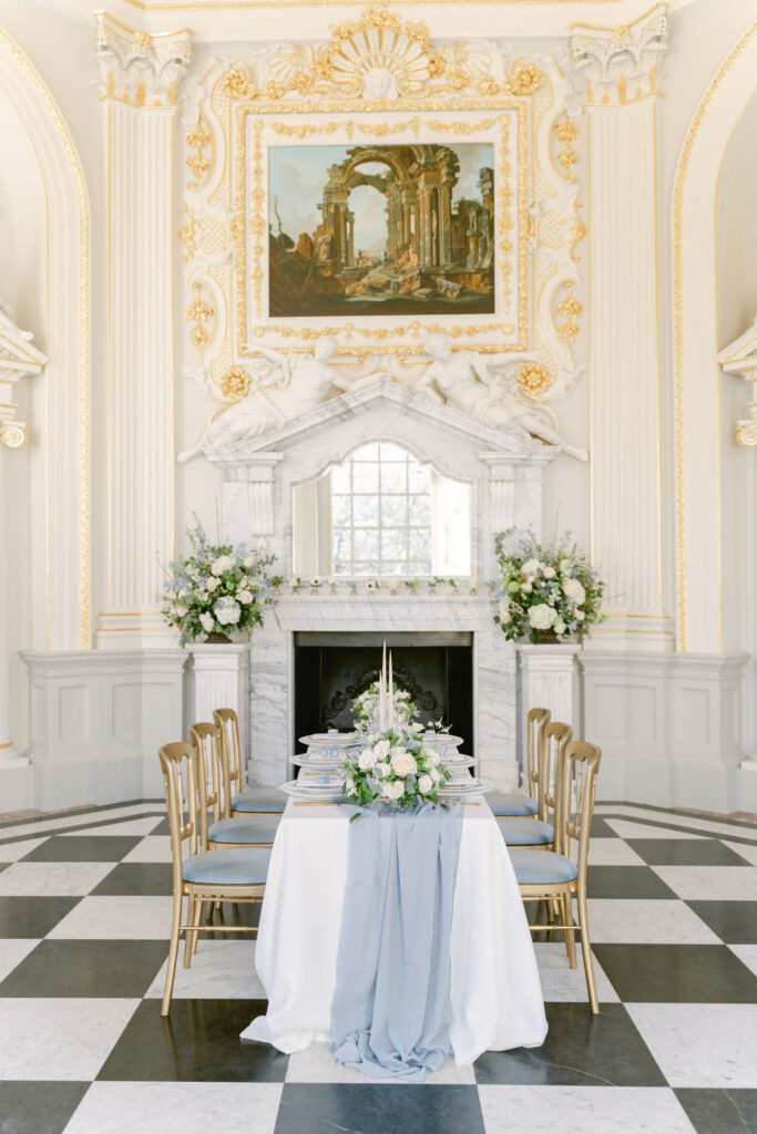 A fine art style wedding breakfast table in white linen with a blue table runner, green and white flower arrangement, set on a black and white checkered floor in the Baroque Octagon Room at Orleans House Gallery.