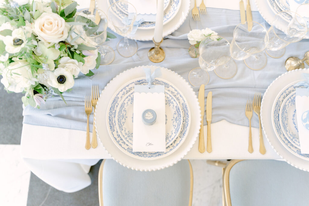 An overhead view of the fine art wedding tablescape featuring blue and white floral crockery on white charger plates, white tablecloth, blue table runner, gold cutlery, gold-rimmed glassware, brass candlesticks with ivory candles and seasonal winter flowers.