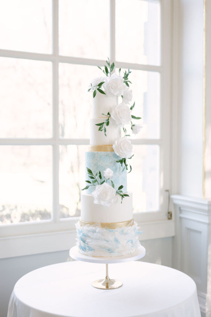 The 5-tiered fine art wedding cake set on a round table against a large window. The cake features white sugar roses and has blue and white tiers.