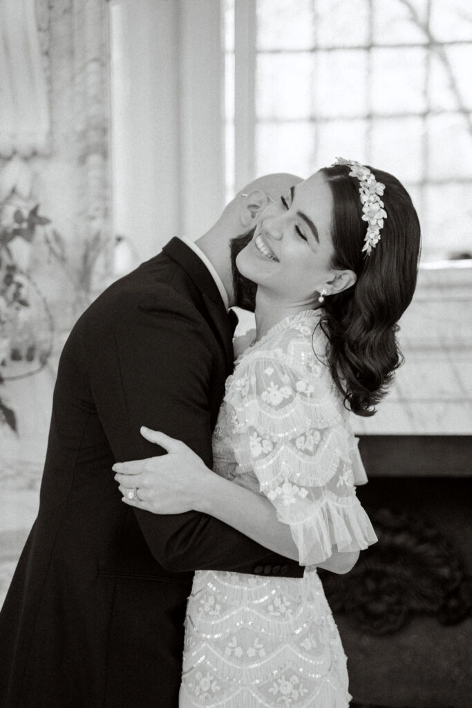 A black and white photograph of the groom kissing the brides neck in the reception room.